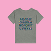 Little Gardeners Grey ABC Toddler T-Shirt featuring a colorful garden-themed ABC illustration on a soft cotton tee on a pink background