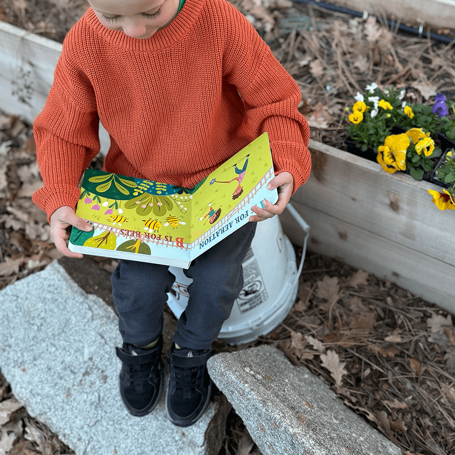 Child in an orange sweater reading a colorful book outdoors with flowers and a planter in the background.